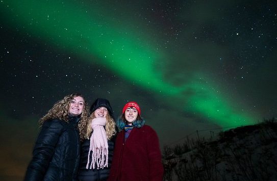 Three women standing below the Aurora Borealis