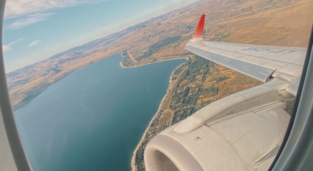 A picture of a destination from inside of a plane, cropped by plane window. Picture features a body of water and wing of plane.