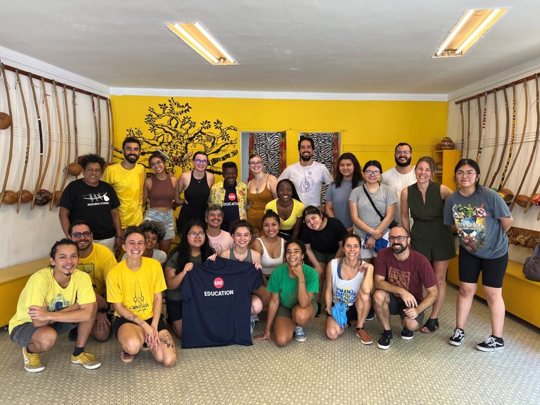 UIC students on the Education, Globalization and the Child in Brazil program pose for a group photo in a room that is painted yellow. A student holds up a shirt that says UIC Education.